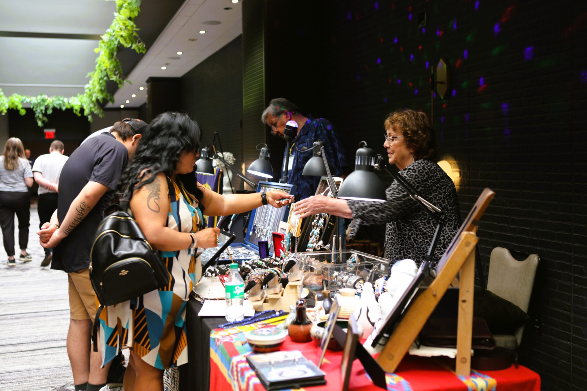 Woman purchasing jewelry from a vendor at an indoor vendor booth with various handmade accessories displayed.