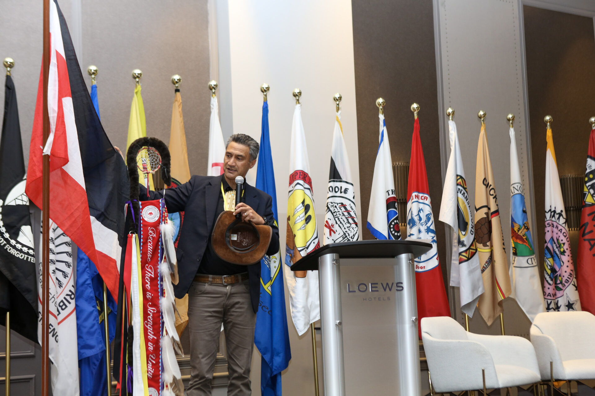 Man holding a hat and speaking into a microphone in front of multiple brightly colored flags on stage at a conference venue.