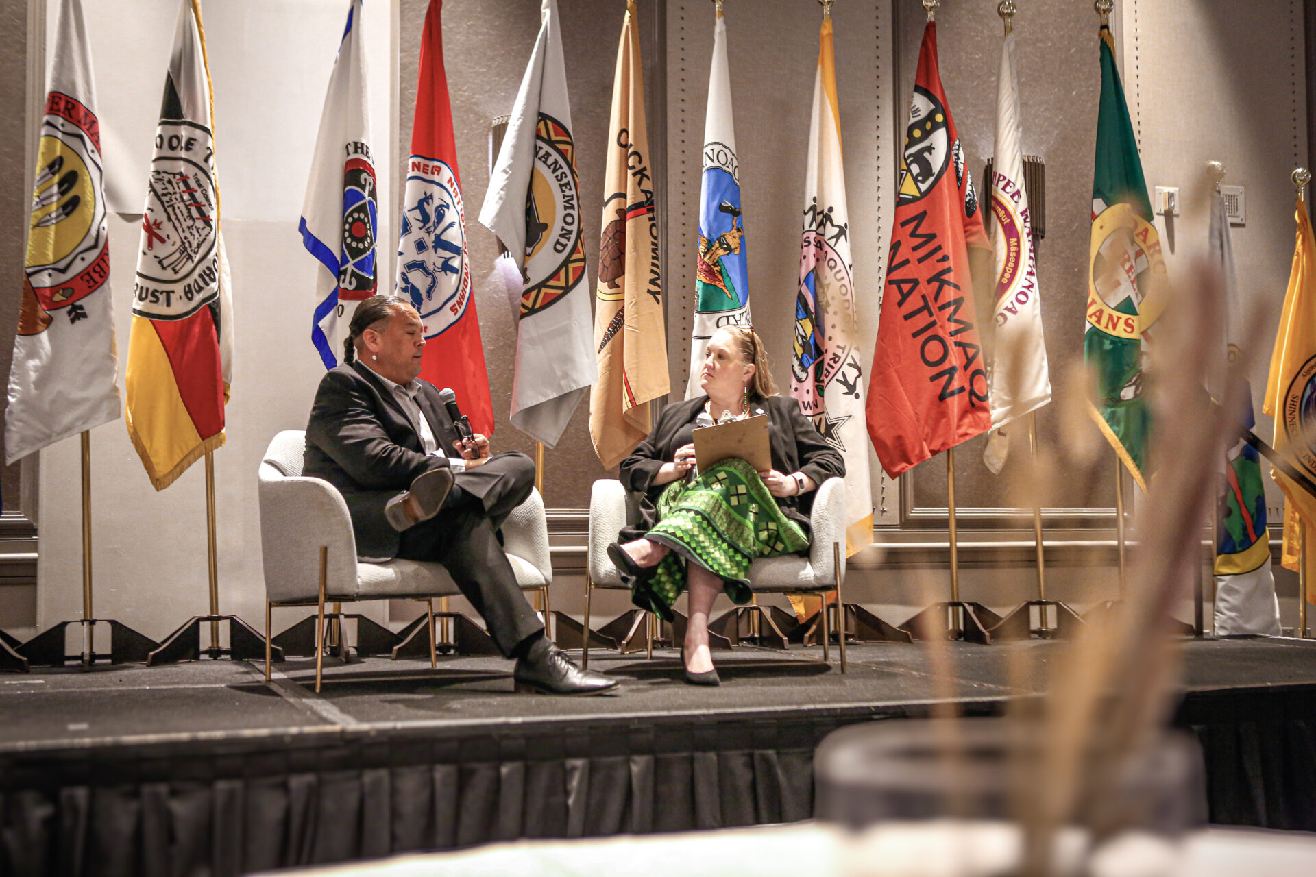 Two people seated on stage engaged in conversation with multiple brightly colored Tribal Nation flags behind them.