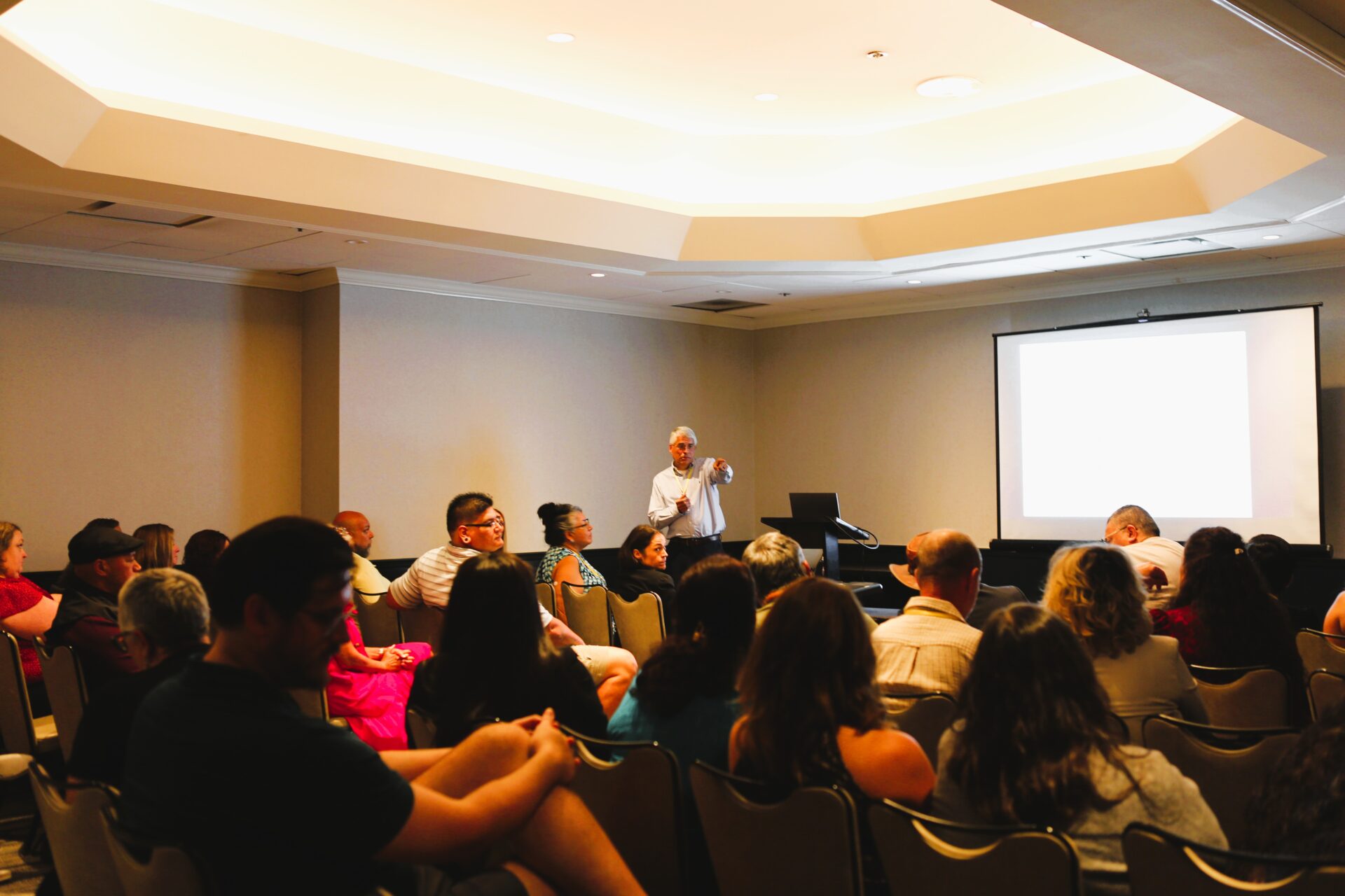 Speaker addressing a seated audience in a conference room with a projection screen behind him.