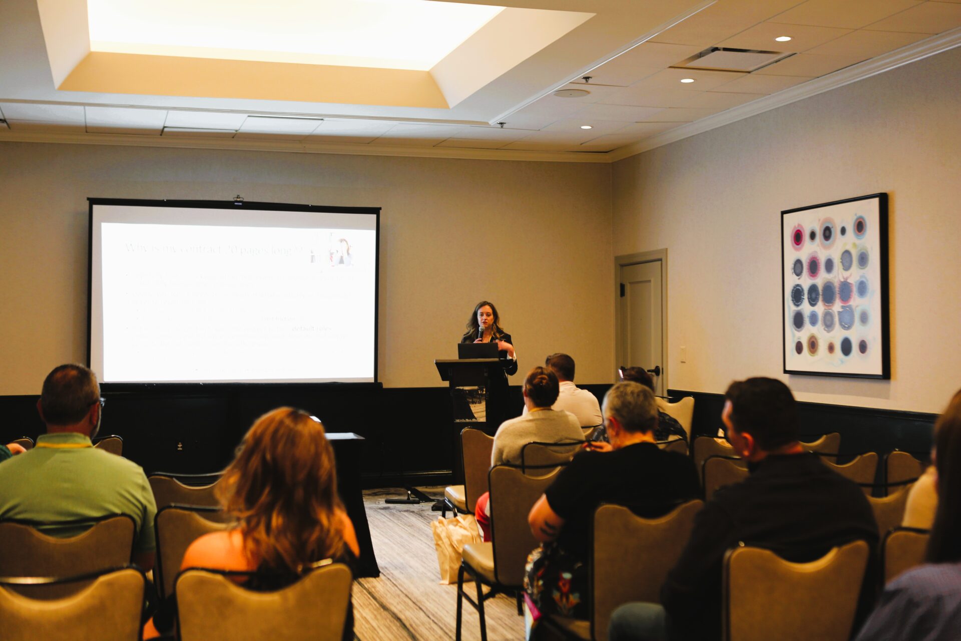 Woman presenting information to an audience in a conference room with a projection screen behind her.