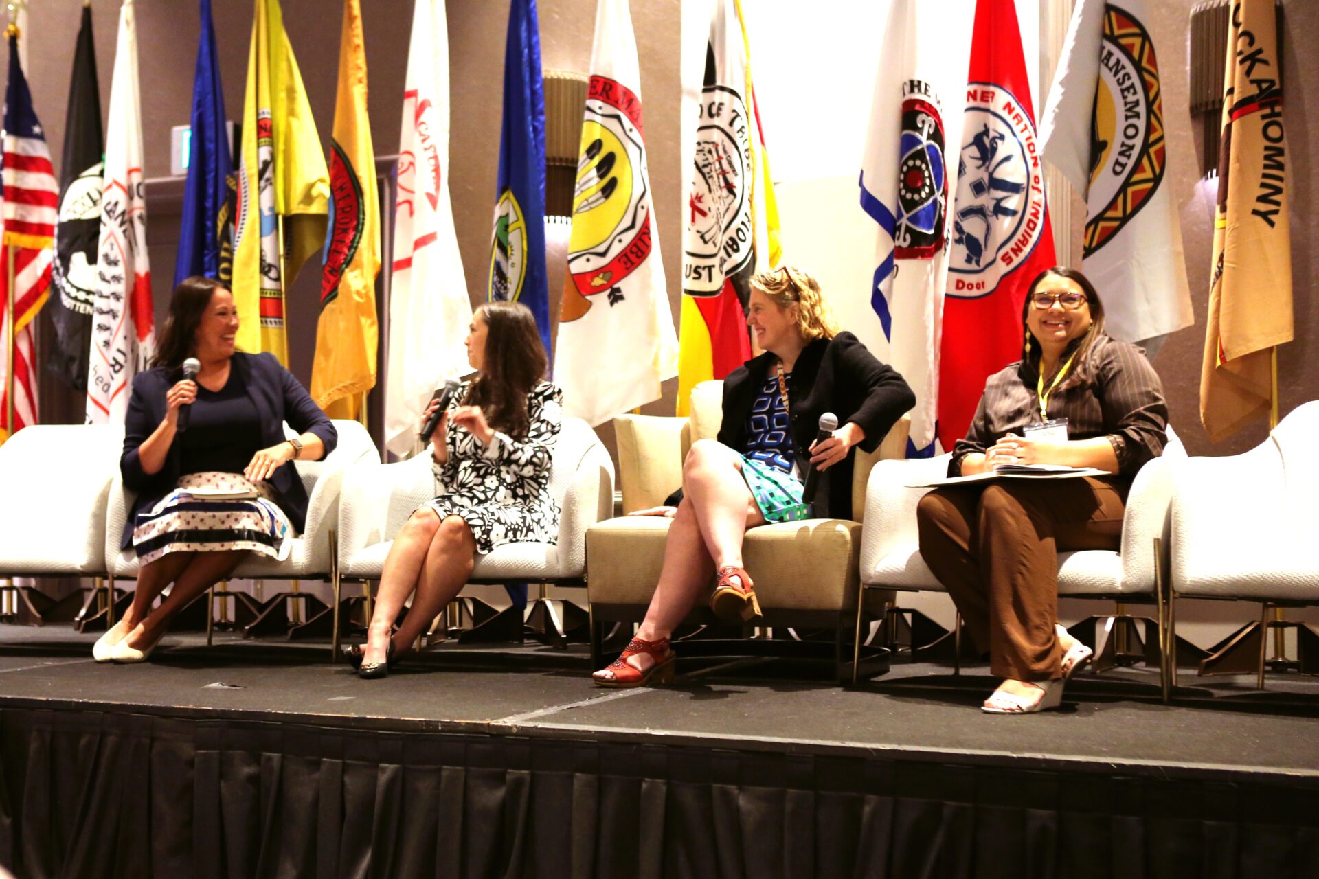 Four women seated on stage engaged in a panel discussion with various brightly colored flags displayed behind them.