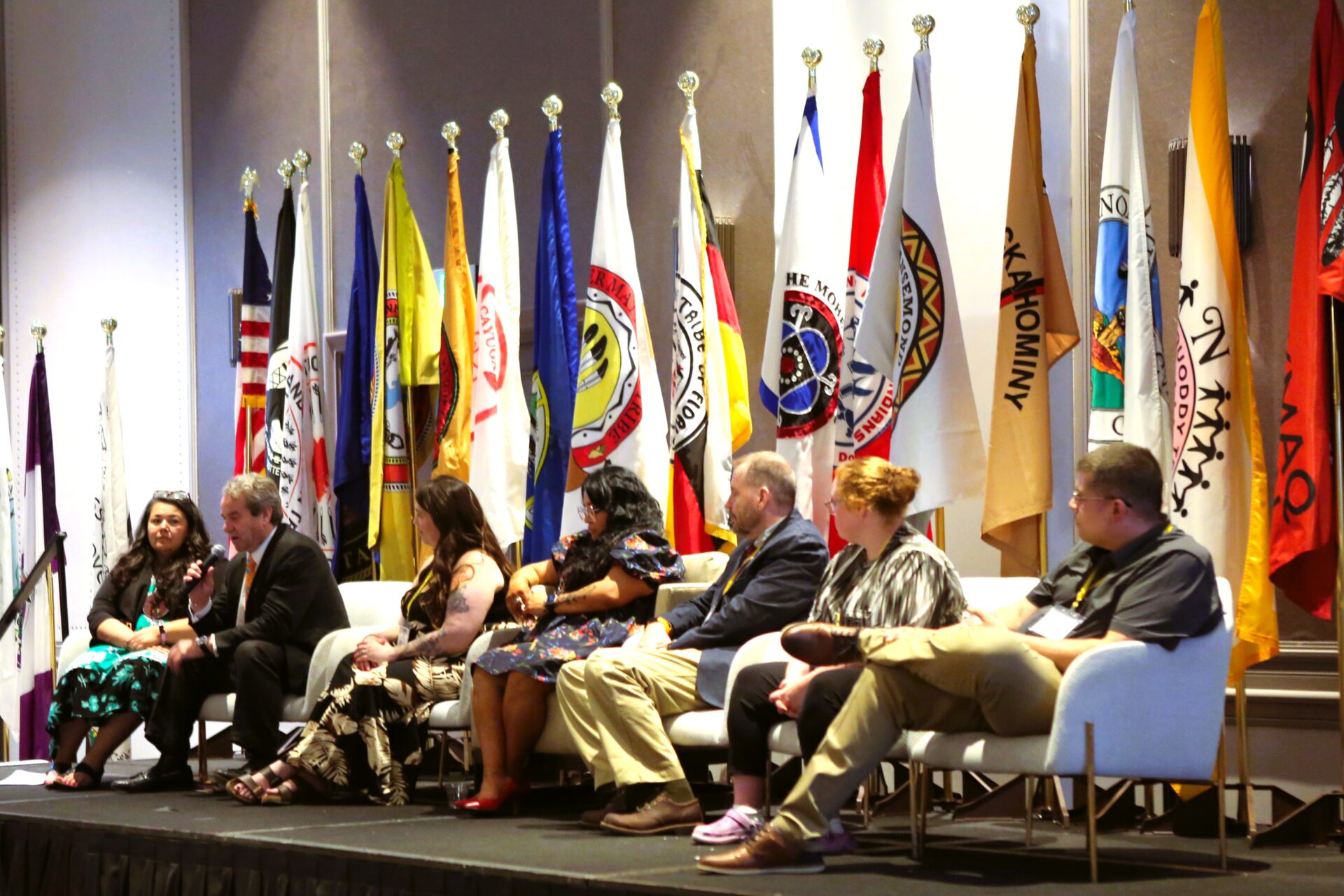 Panel of seven speakers seated on stage with multiple colorful flags behind them at a formal event.