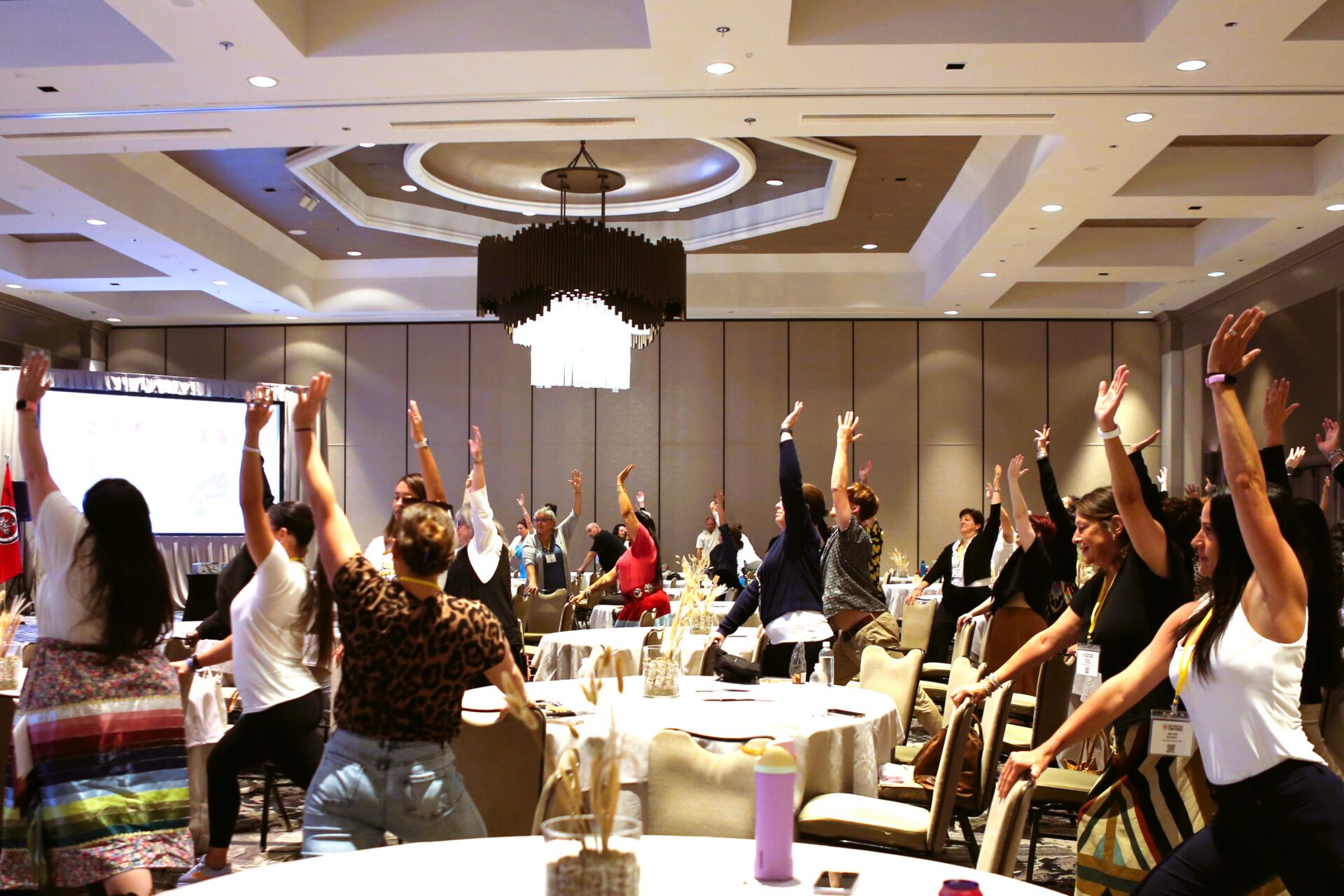 Group of people in a large conference room raising their arms during a group yoga activity.