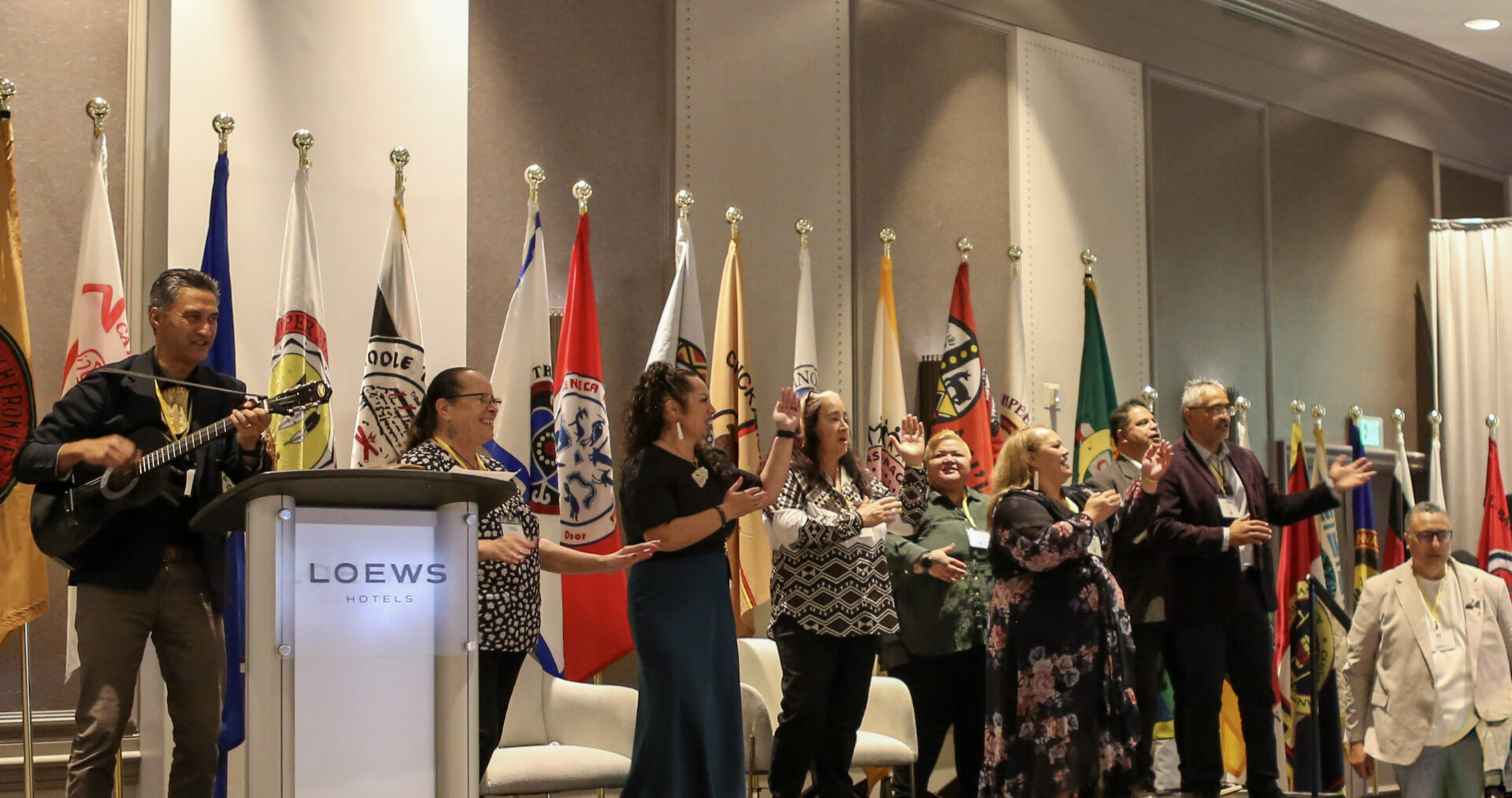 Group of people on stage in a large conference room, some singing and one playing guitar, with various flags in the background.