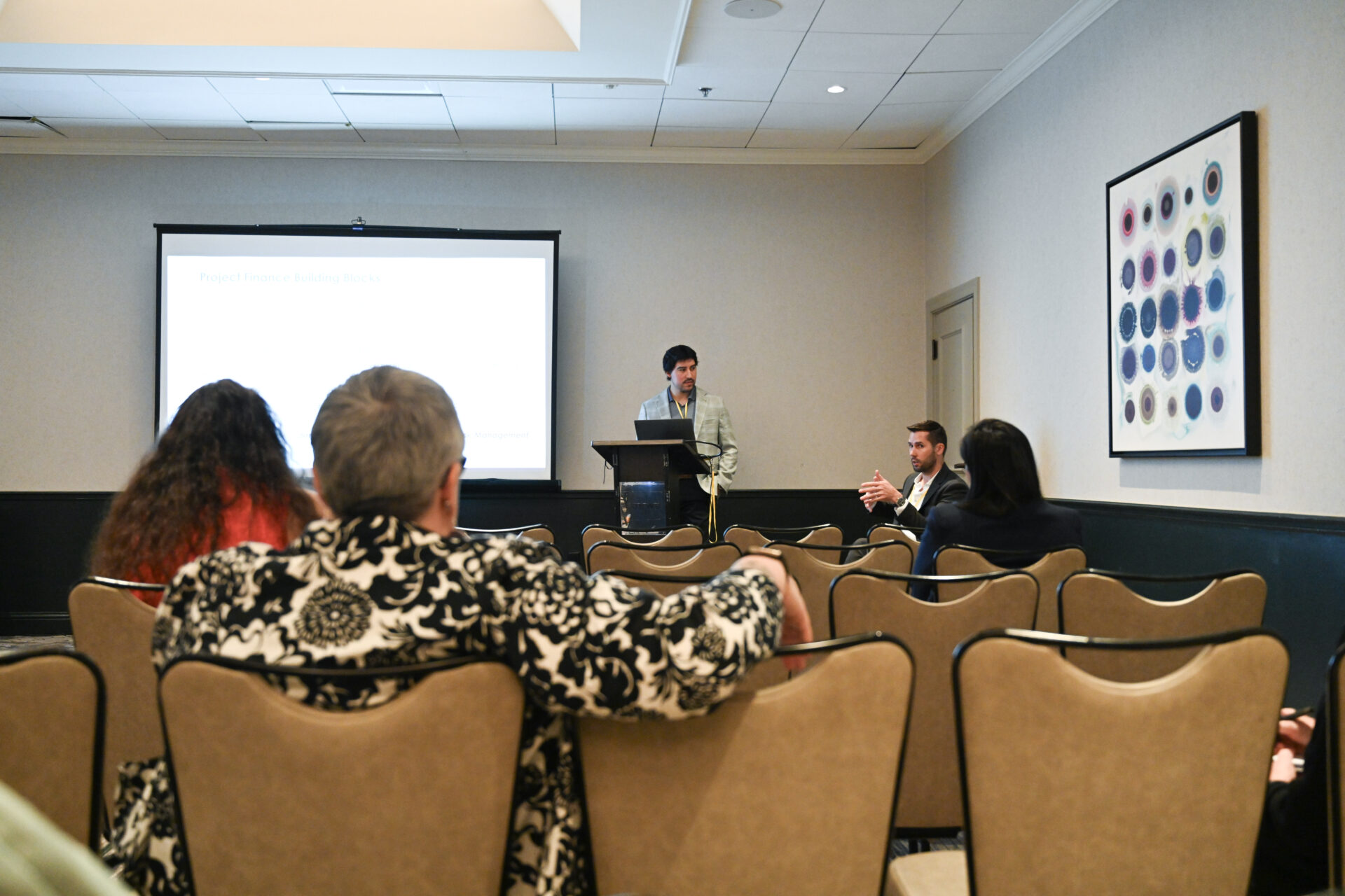 A group attending a presentation in a conference room with a speaker at the podium and a man engaging with the audience.