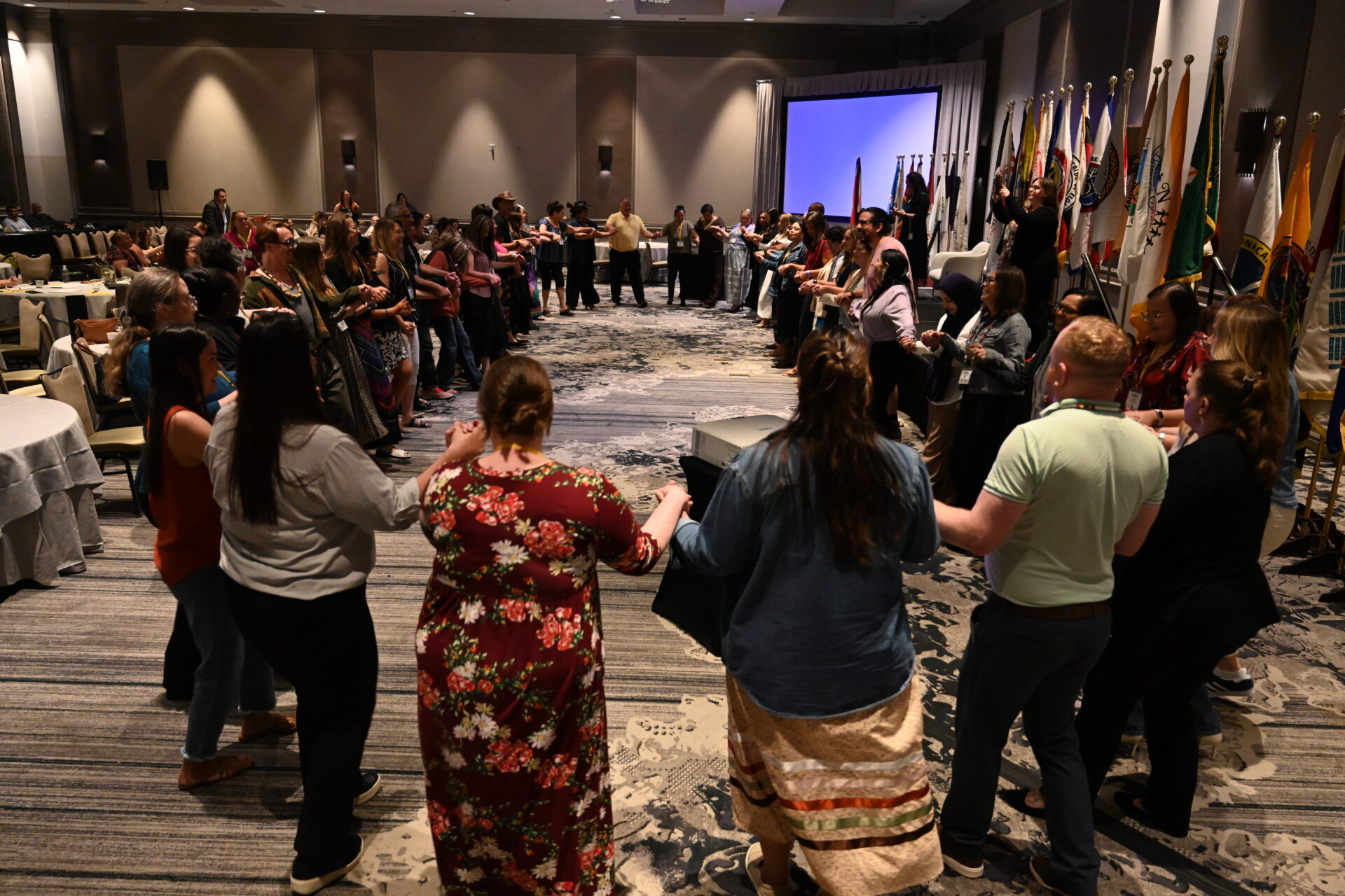A large group of people standing in a conference room holding hands in a circle during a social round dance.