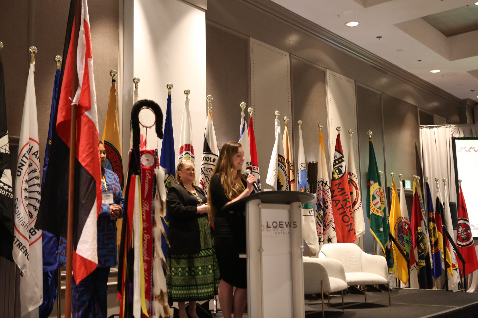 Woman speaking into a microphone in front of multiple brightly colored flags on stage at a conference venue.